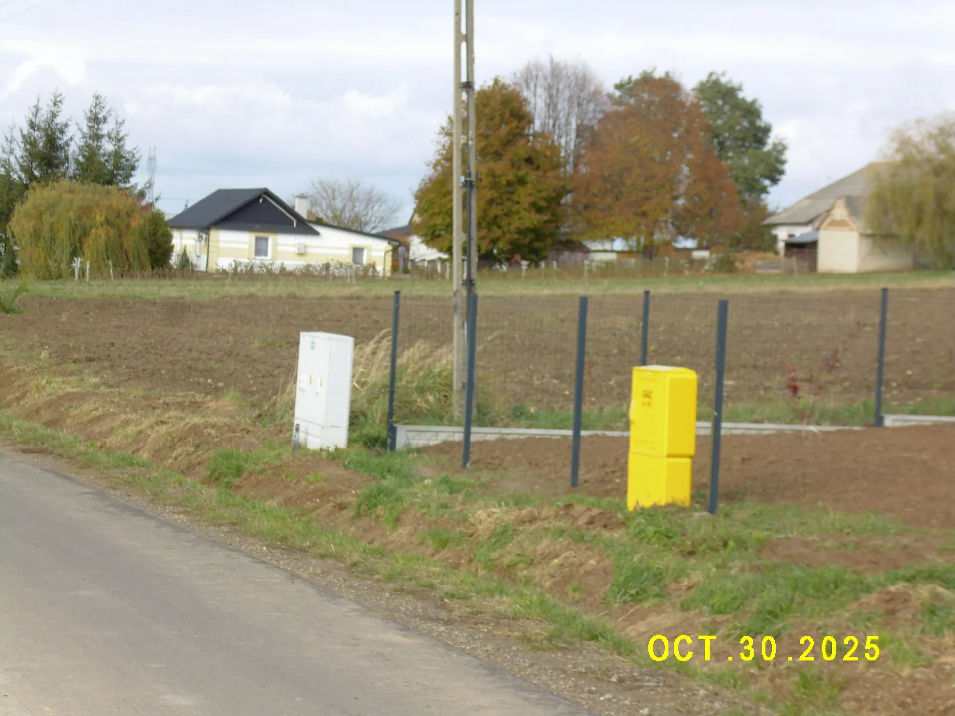 Krajobraz wiejski z asfaltową drogą w lewym dolnym rogu. Przy drodze znajdują się dwie szafki elektryczne: jedna biała i jedna żółta, oddzielone od pola metalowym ogrodzeniem z siatki. W tle widoczny jest biały dom z ciemnym dachem, jesienne drzewa o żółtych i pomarańczowych liściach oraz słup energetyczny. W prawym dolnym rogu znajduje się żółty napis z datą: OCT.30.2025