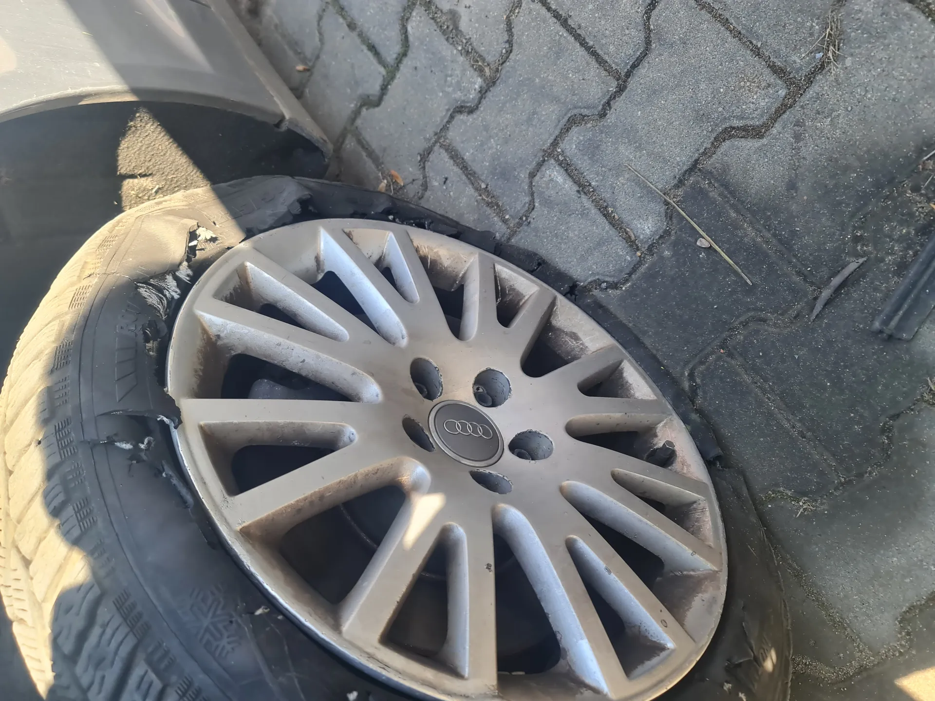 Zdjęcie przedstawia uszkodzone koło samochodowe z felgą marki Audi (cztery połączone pierścienie w środku). Opona jest pęknięta i zdeformowana, zwłaszcza po lewej stronie. Koło leży na chodniku z betonowych płyt ułożonych w geometryczny wzór. W tle widoczne są fragmenty asfaltu oraz drobne przedmioty, takie jak patyk. Oświetlenie wskazuje na słoneczny dzień, z cieniem padającym z lewej strony obiektu.