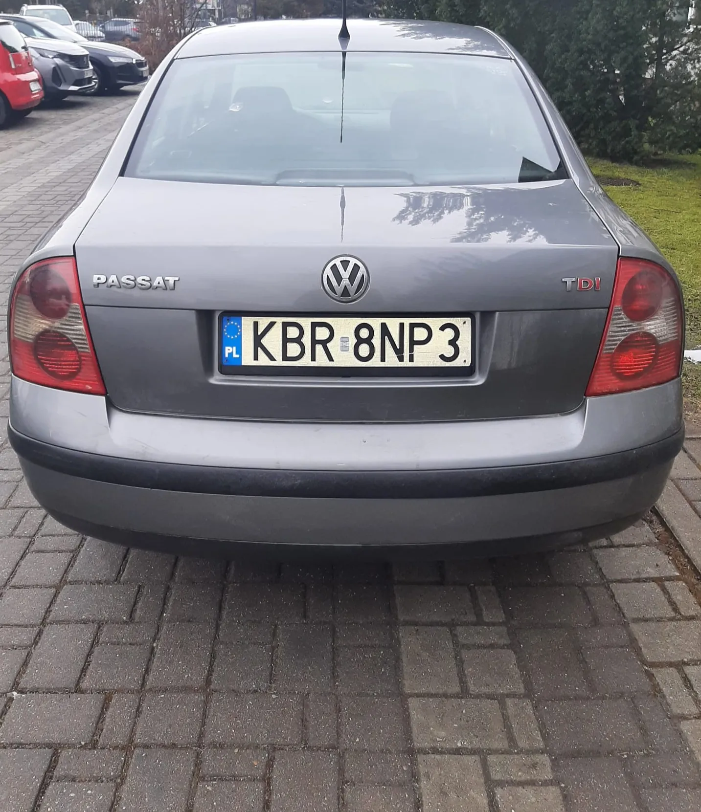Tył szarego samochodu Volkswagen Passat TDI zaparkowanego na brukowanej powierzchni. Widoczna tablica rejestracyjna z polskim kodem PL i numerem KBR 8NP3. Po lewej stronie napis PASSAT, po prawej TDI, a w centrum logo Volkswagena. W tle inne zaparkowane samochody oraz zieleń.