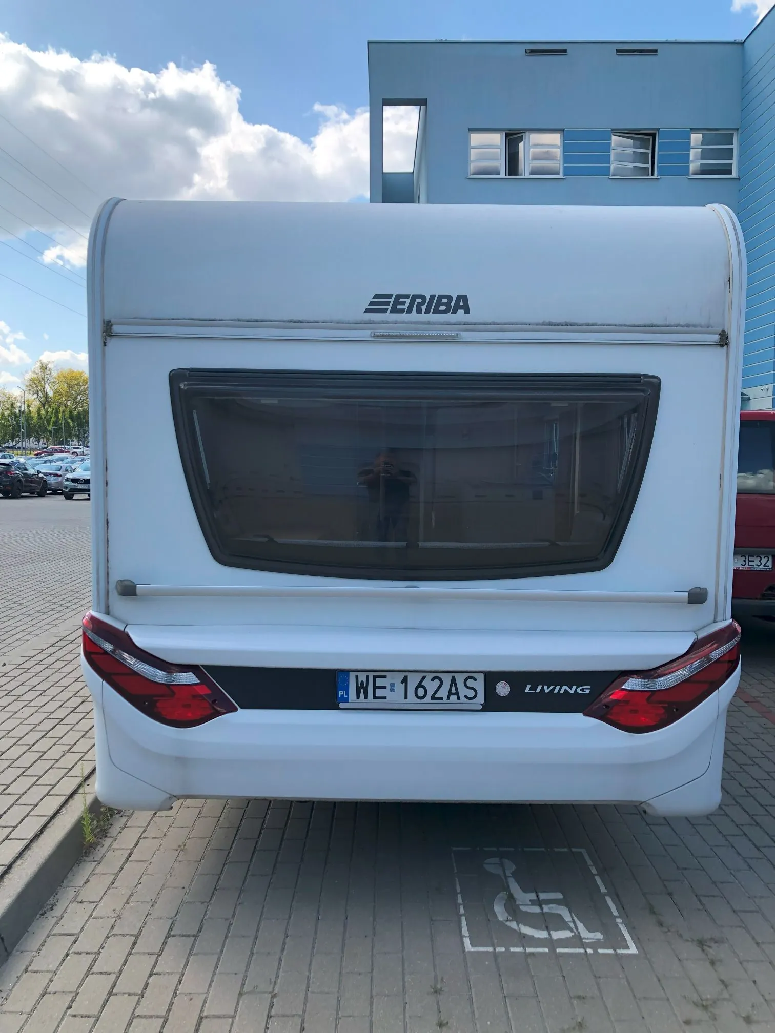 Tylny widok białego kampera marki Eriba, model Living. Pojazd posiada ciemną szybę tylną, czerwone lampy tylne oraz polską tablicę rejestracyjną o numerze WE 162AS. Kamper zaparkowany jest na kostce brukowej, na miejscu wyznaczonym dla osób z niepełnosprawnością, co sygnalizuje biały symbol wózka inwalidzkiego na podłożu. W tle widoczny jest niebieski budynek biurowy oraz fragmenty innych samochodów pod jasnym, częściowo zachmurzonym niebem.