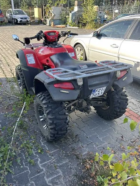 Czerwony quad zaparkowany na kostce brukowej obok srebrnego samochodu osobowego. Pojazd posiada szary bagażnik z tyłu oraz tablicę rejestracyjną o numerze FZI 3051. W tle widoczny jest inny srebrny samochód oraz fragmenty zabudowań i drzew.