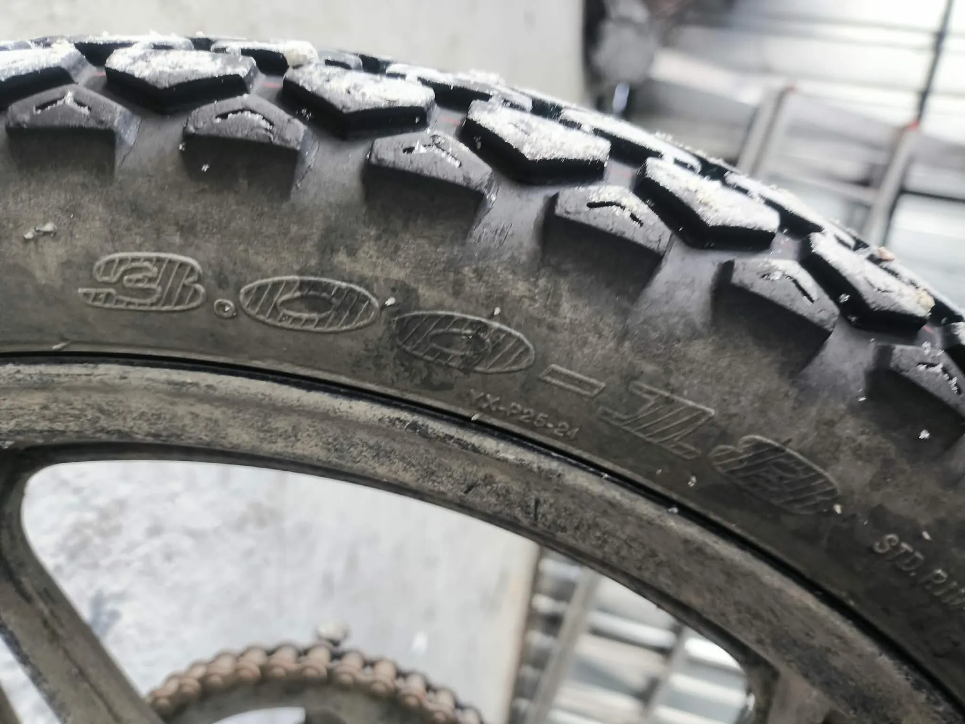 Zbliżenie na oponę z widocznym wzorem gąsienicy i wytłoczonymi napisami: 90/90-21, STD.R1 oraz logo CST. Tło przedstawia przemysłowe pomieszczenie z metalowymi konstrukcjami i łańcuchem. Opona jest zabrudzona, z białymi plamami na gąsienicy.