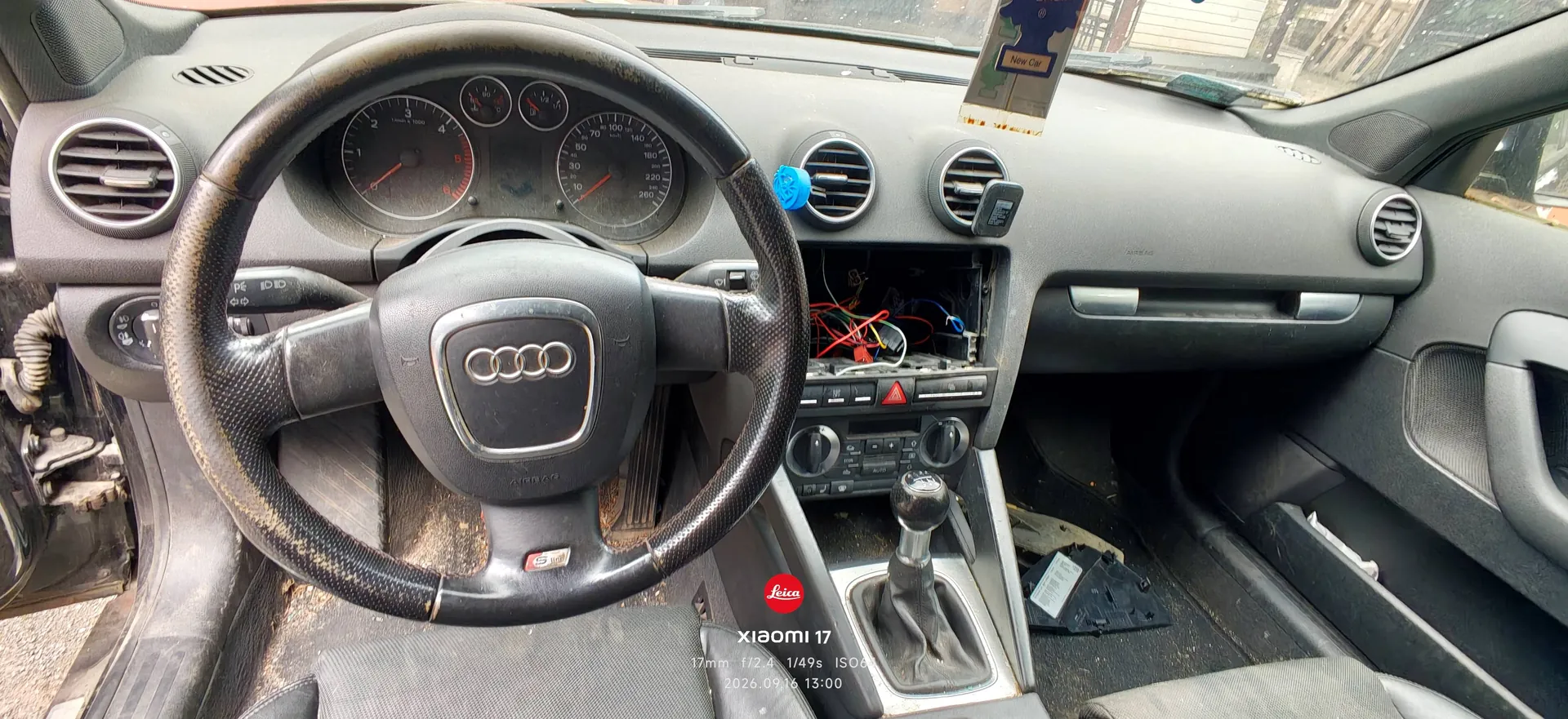 Wnętrze samochodu marki Audi z perspektywy kierowcy. Widoczna jest czarna kierownica z logo Audi w centrum, deska rozdzielcza z licznikiem prędkości i obrotomierzem oraz otwory nawiewów. W centralnej części deski rozdzielczej brakuje radia, odsłaniając kolorowe przewody elektryczne. Poniżej znajduje się panel sterowania klimatyzacją i lewarek zmiany biegów. Na podszybiu zamocowany jest uchwyt na telefon. W stopie pasażera leżą ciemne przedmioty.
