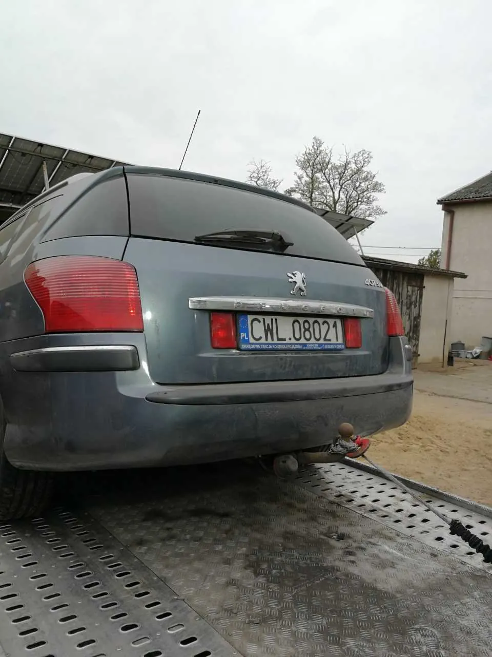 Tył szarego samochodu marki Peugeot 407 z widocznym logo lwa i napisem "PEUGEOT". Tablica rejestracyjna PL CWL.08021 z nieczytelnym napisem poniżej. Samochód jest przytwierdzony do metalowej platformy za pomocą haków holowniczych. W tle budynki, drzewa bez liści i pochmurne niebo. Podłoga platformy ma wzór rombów i otwory.