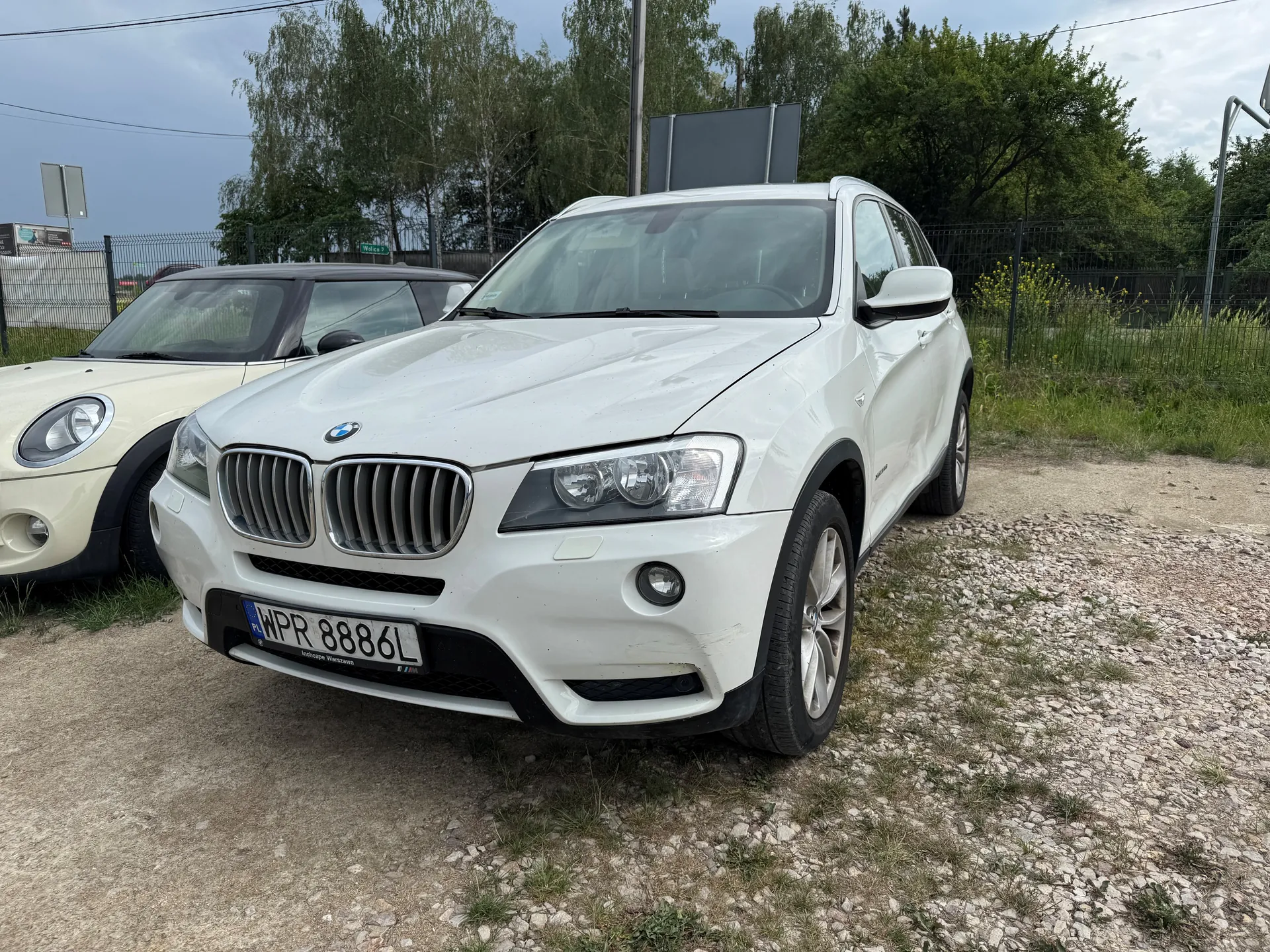 Biały samochód BMW z tablicą rejestracyjną WPR 8886L zaparkowany na żwirowym placu. Obok widoczny beżowy samochód Mini Cooper. W tle drzewa, ogrodzenie, znaki drogowe oraz pochmurne niebo. Na tablicy rejestracyjnej widoczny napis 'PL' oraz 'WPR 8886L'.