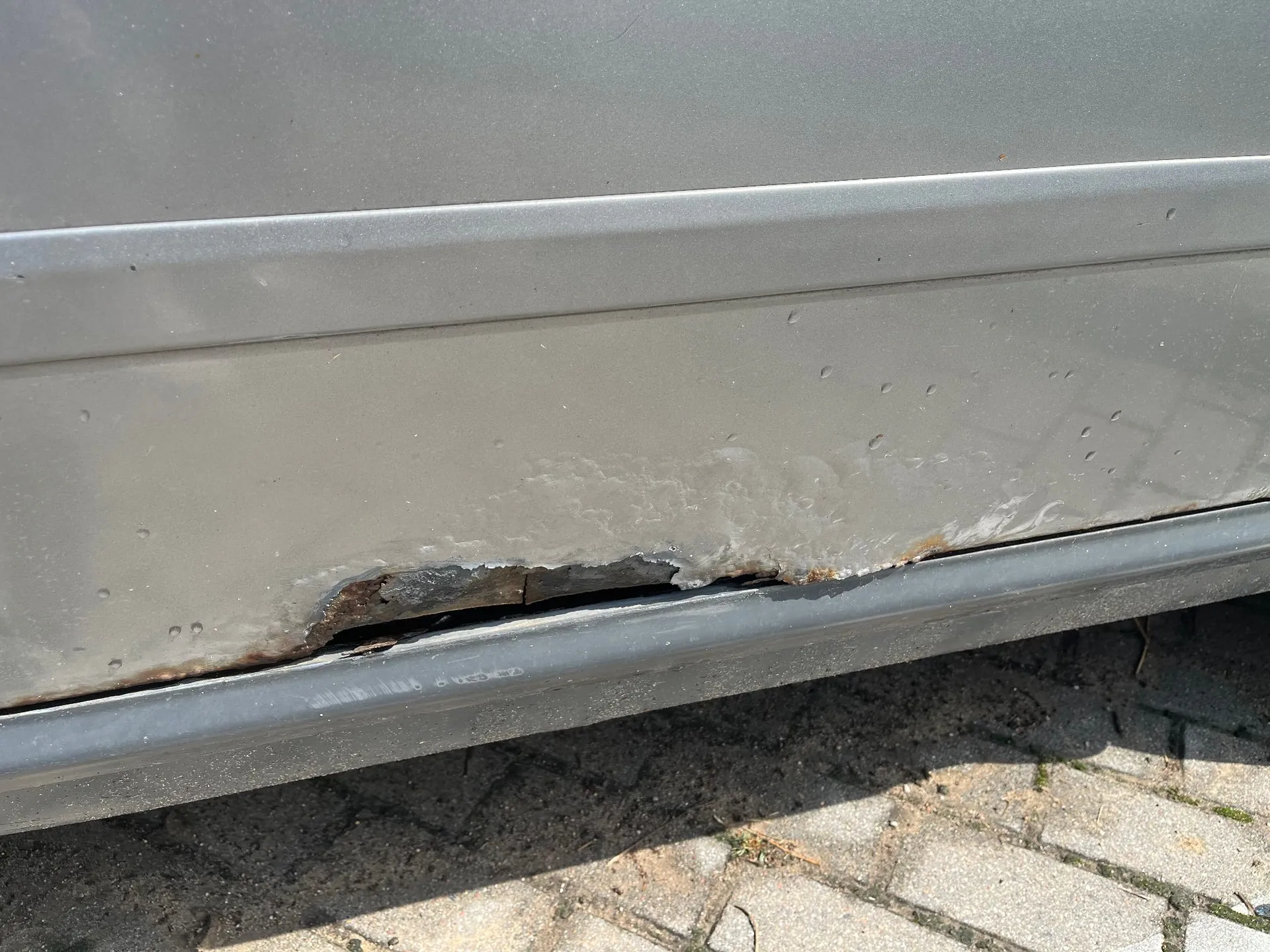 Zbliżenie na dolną część srebrnego nadwozia samochodu, w której widoczne jest znaczne uszkodzenie blachy. W dolnej krawędzi progu znajduje się głęboka rdza i wyrwa w metalu, odsłaniająca ciemne wnętrze konstrukcji. Powierzchnia lakieru wokół uszkodzenia jest zmatowiała i posiada drobne wgniecenia. Samochód stoi na kostce brukowej.