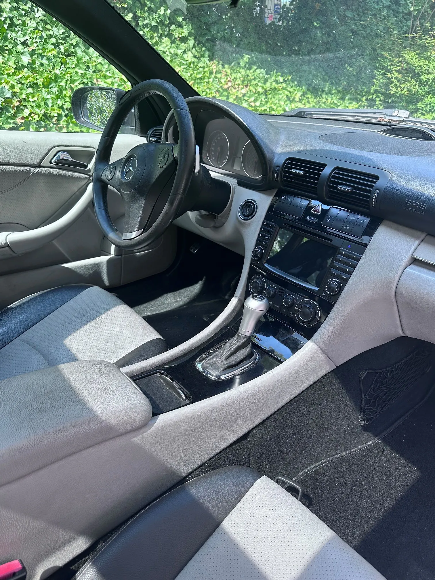 Wnętrze samochodu marki Mercedes-Benz widziane z perspektywy pasażera. Widoczna jest szara tapicerka skórzana, czarna kierownica z logo marki, deska rozdzielcza z licznikiem, systemem audio z ekranem oraz lewarem zmiany biegów. Przez przednią szybę i okno widać zielone liście drzew.