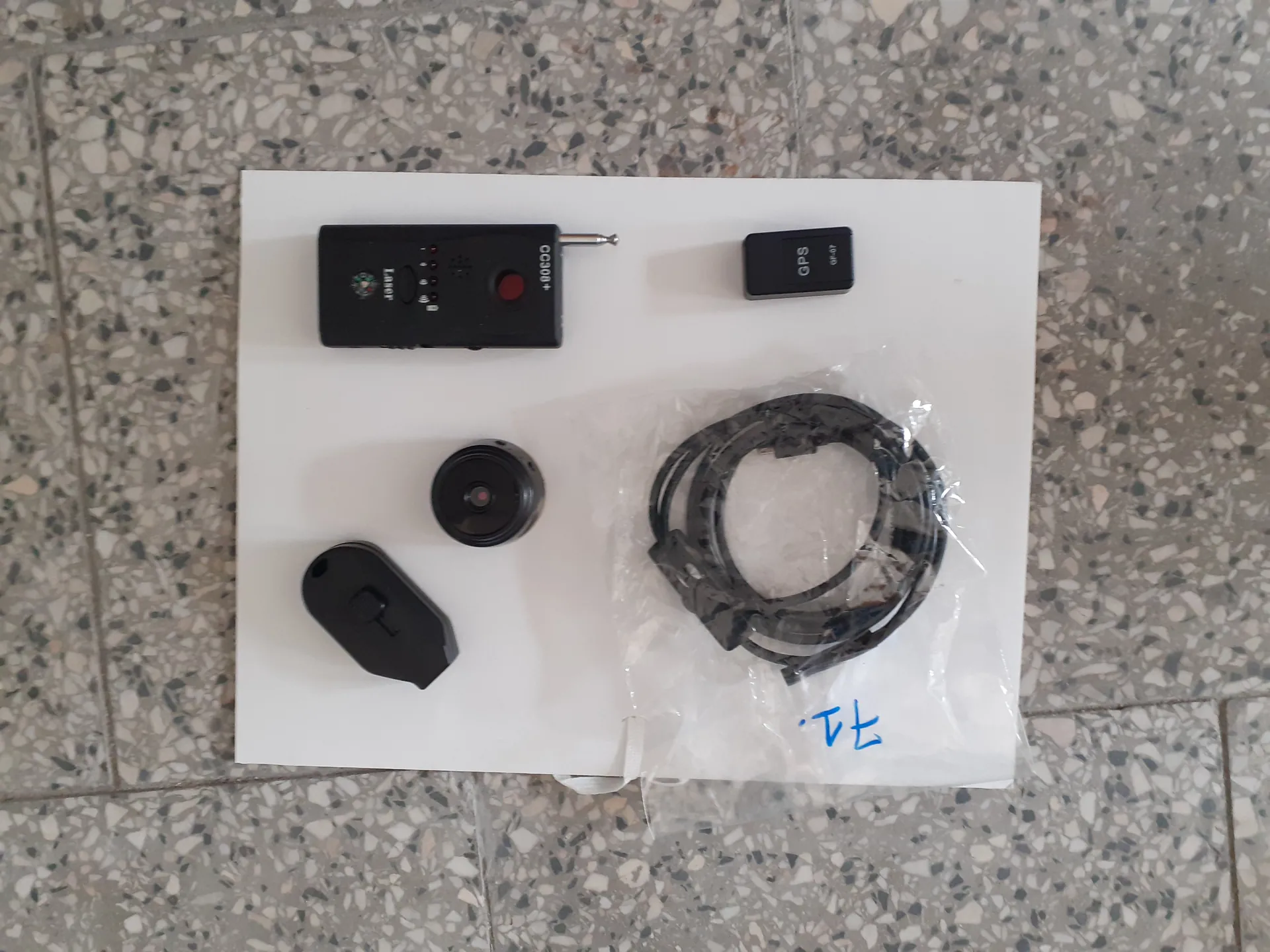 Na białym podłożu umieszczono cztery czarne urządzenia elektroniczne i zwinięty kabel w przezroczystej torbie. Po lewej stronie znajduje się prostokątny aparat z czerwonym przyciskiem, napisem OC300+ oraz małym logo. Obok niego, po prawej, mały prostokątny moduł z napisem GPS. Poniżej widoczny jest okrągły obiektyw kamery oraz czarny, owalny element z przyciskiem. W przezroczystej torbie znajduje się czarny kabel, a na niej niebieskim markerem zapisano T2. Tło stanowi podłoga z kafelkami w kolorze szaro-białym z czarnymi plamkami.