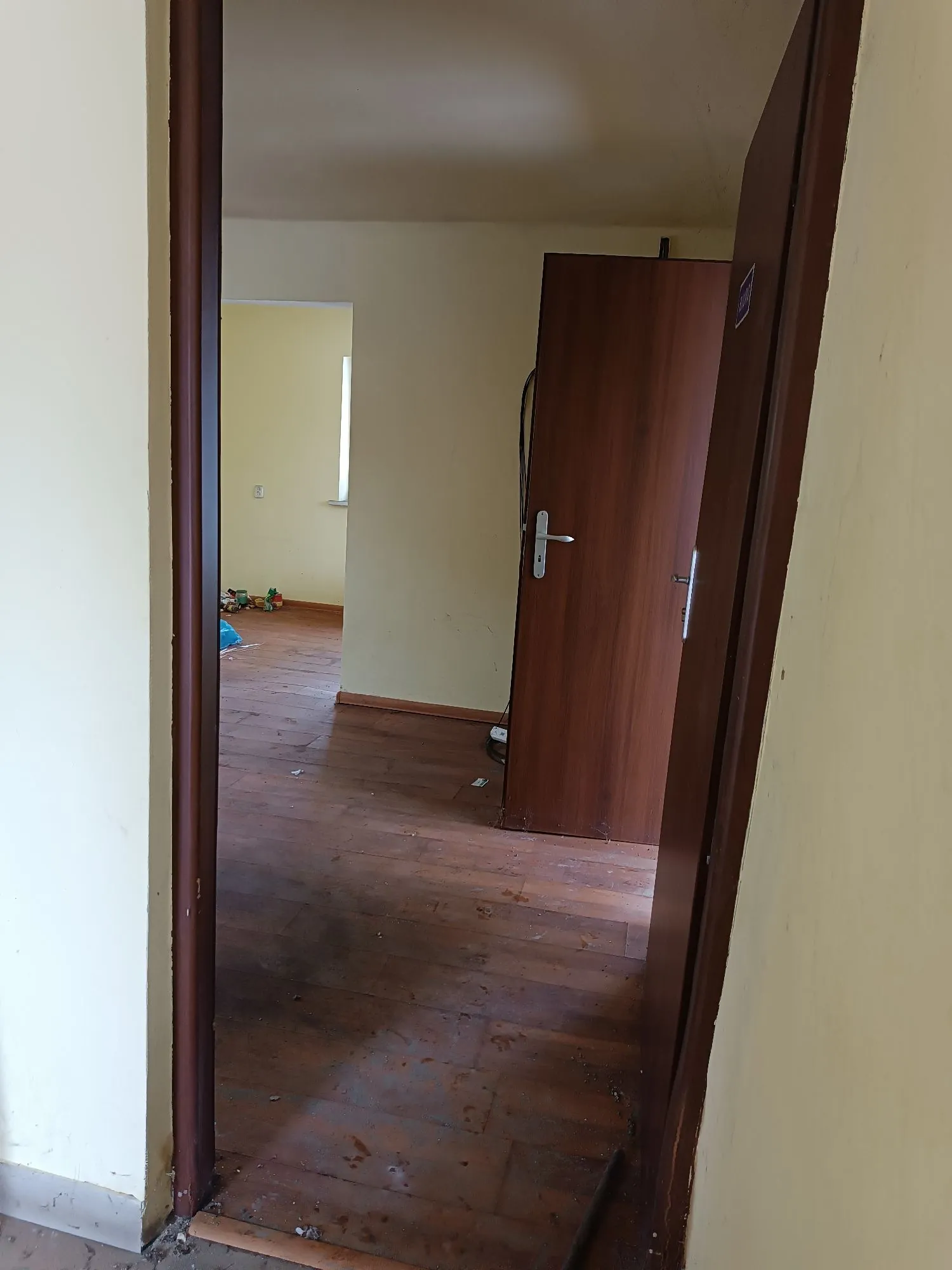 Widok z korytarza przez otwarte drzwi do pokoju z drewnianą podłogą i jasnożółtymi ścianami. Wewnątrz pokoju widoczne są kolejne otwarte drzwi prowadzące do innego pomieszczenia oraz fragmenty zabawek na podłodze w oddali.