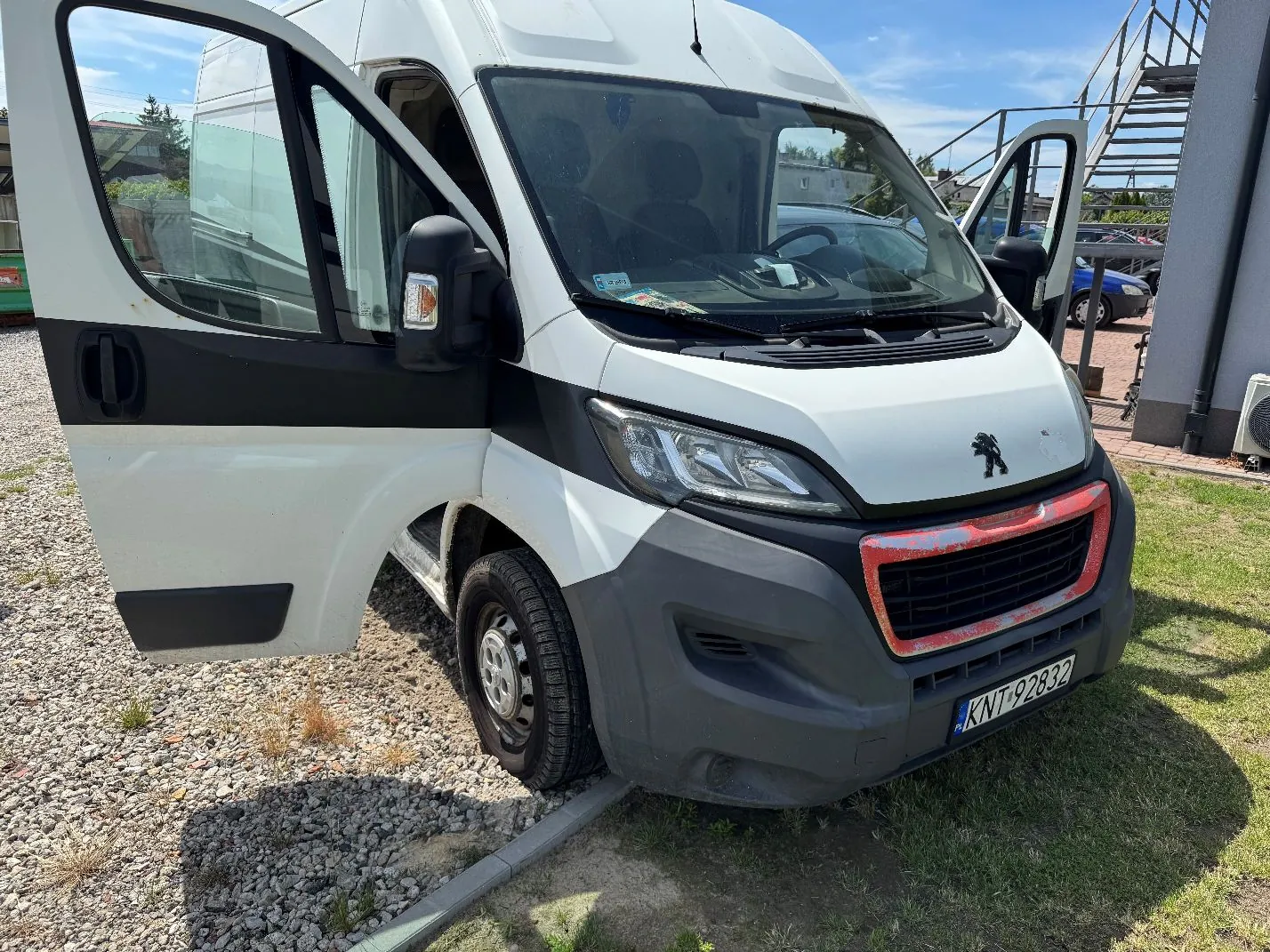 Biały samochód dostawczy marki Peugeot z otwartymi przednimi drzwiami, zaparkowany na żwirze i trawie. Pojazd ma szary zderzak z czerwonymi śladami uszkodzeń na grillu. Tablica rejestracyjna ma numer KNT 92832.