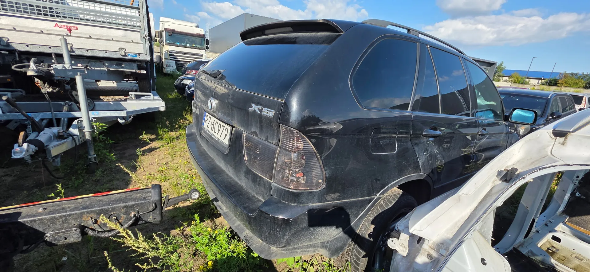 Czarny samochód BMW X5 zaparkowany na trawiastym terenie. Widoczny jest tył pojazdu z tablicą rejestracyjną PZ 6C979. W tle znajdują się inne samochody, biała naczepa ciężarówki oraz fragmenty metalowych konstrukcji i przyczep. Niebo jest jasnoniebieskie z białymi chmurami.