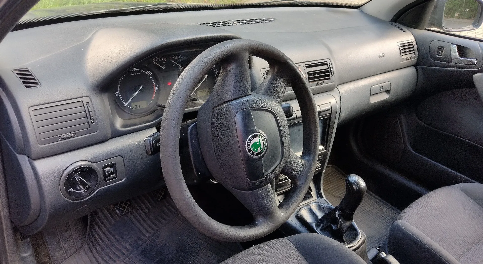 Wnętrze samochodu marki Skoda, widok z perspektywy kierowcy. Widoczna jest czarna kierownica z logo marki w centrum, deska rozdzielcza z analogowymi wskaźnikami prędkości i obrotów, kratki nawiewów oraz lewarek zmiany biegów. Fotele są obite ciemnoszarą tkaniną.