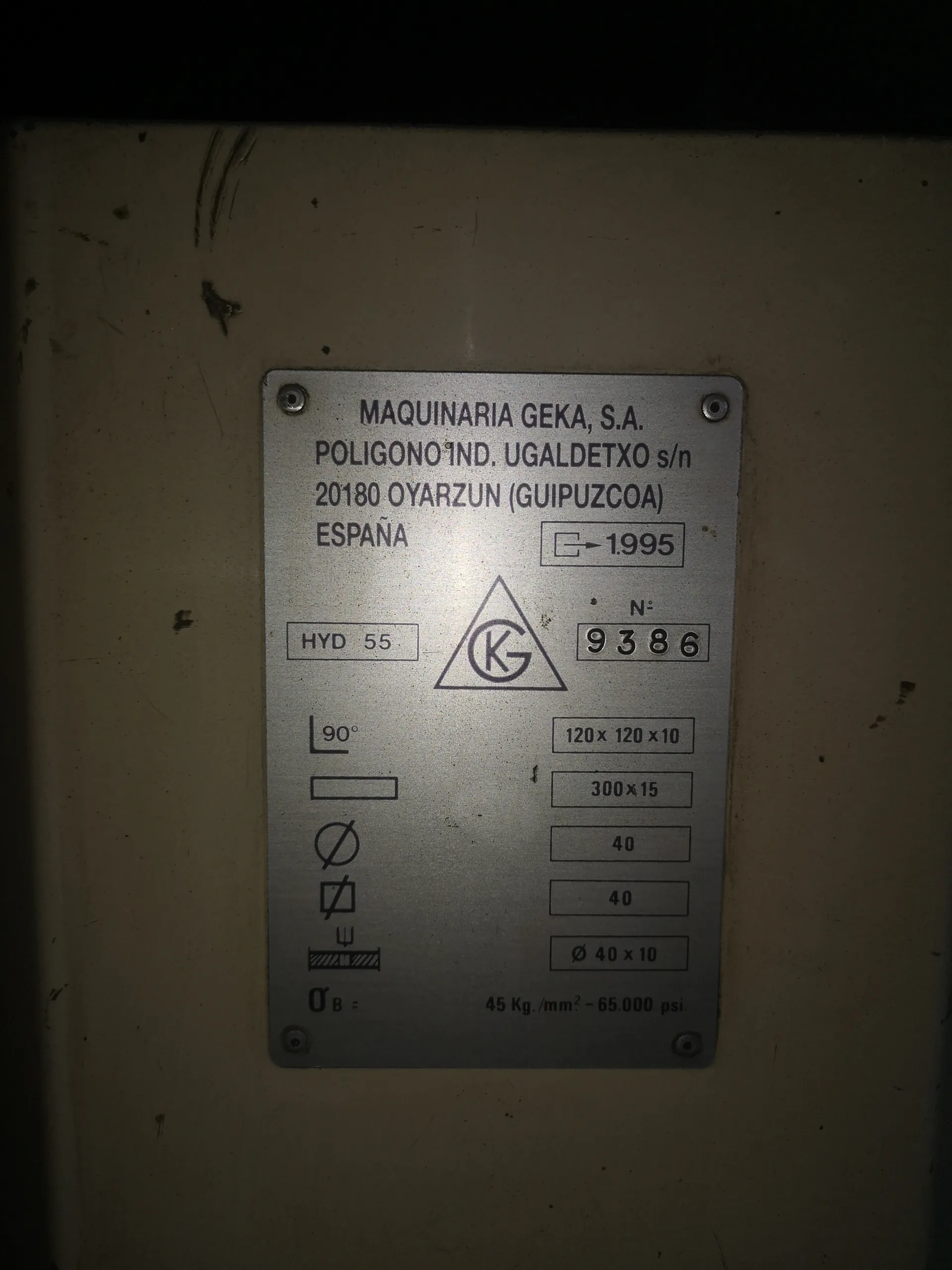 Metalowa tabliczka zabezpieczona śrubami, zawierająca tekst i symbole. Widoczny napis: MAQUINARIA GEKA, S.A. POLIGONO IND. UGALDETXO s/n 20180 OYARZUN (GUIPUZCOA) ESPAÑA. Obok prostokąt z napisem 1995. Poniżej: HYD 55, trójkąt z literami KG, numer N° 9386. Techniczne dane: 120 x 120 x 10, 300 x 15, 40, 40, Ø 40 x 10. Po lewej stronie symbole: 90°, pionowa kreska, zakazany okrąg, kwadrat z przekreśloną linią, symbol podparcia. Na dole: σ_B = 45 Kg./mm² - 65.000 psi. Tło stanowi metalowa powierzchnia z widocznymi zadrapaniami.