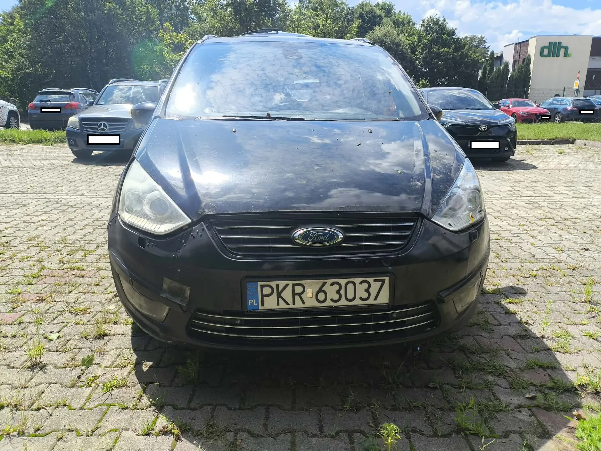 Czarny samochód marki Ford widziany od przodu, zaparkowany na kostce brukowej z trawą w szczelinach. Pojazd ma polską tablicę rejestracyjną o numerze PKR 63037. W tle widoczne są inne zaparkowane samochody oraz budynek z napisem 'dlh'.