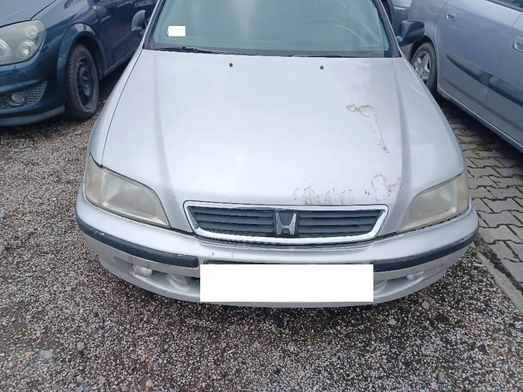Srebrny samochód marki Honda widziany od przodu, zaparkowany na żwirowym podłożu. Na masce widoczne są uszkodzenia lakieru i zacieki. Tablica rejestracyjna jest zakryta białym polem. Po lewej stronie widoczny jest fragment ciemnoniebieskiego samochodu, a po prawej fragment srebrnego auta.
