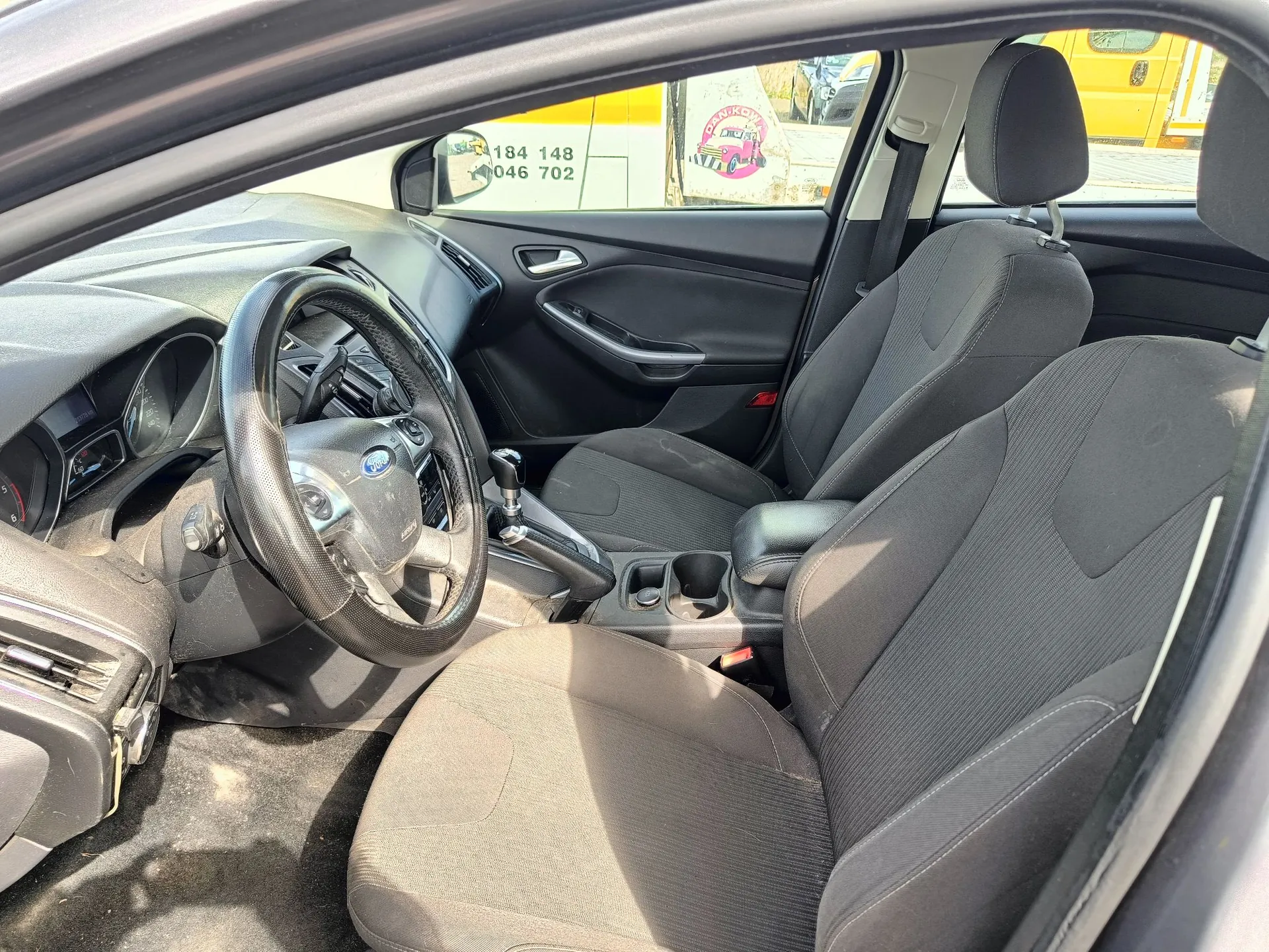 Wnętrze samochodu marki Ford widziane z otwartych drzwi kierowcy. Widoczna jest czarna tapicerka materiałowa na fotelach, czarna kierownica z logo Forda, deska rozdzielcza oraz konsola środkowa z uchwytami na kubki. Przez szybę w tle widoczny jest fragment białego pojazdu z numerami telefonów 184 148 oraz 046 702.