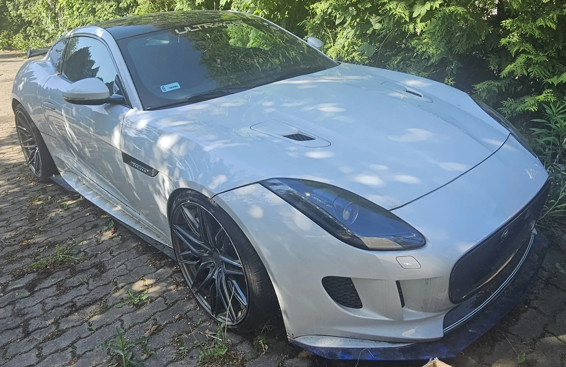 Srebrny samochód sportowy Jaguar F-Type zaparkowany na kostce brukowej obok zielonych krzewów. Auto posiada czarne felgi, czarny dach oraz napis 'ULTRAC' na górnej części przedniej szyby.