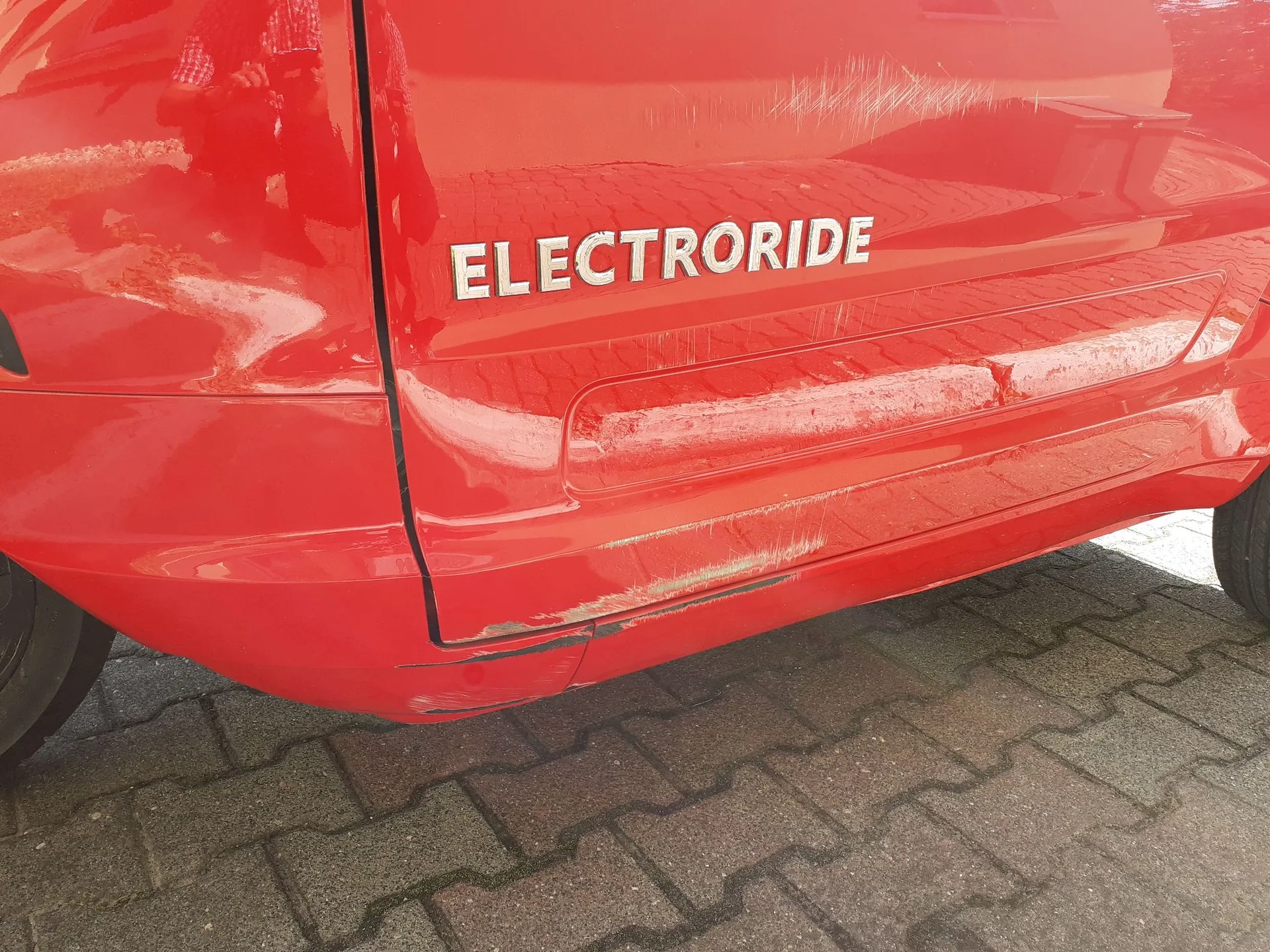 Zbliżenie na tylną część czerwonego samochodu. Na karoserii znajduje się biały napis drukowanymi literami: ELECTRORIDE. Lakier w kilku miejscach jest porysowany. Samochód stoi na kostce brukowej.