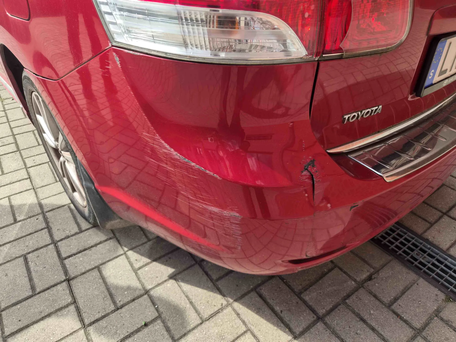 Zbliżenie na tylny, czerwony zderzak samochodu marki Toyota. Na lakierze widoczne są liczne zarysowania oraz wgłębienie w dolnej części zderzaka. W górnej części zdjęcia widoczna jest tylna lampa oraz napis TOYOTA na chromowanej listwie. Samochód zaparkowany jest na kostce brukowej.