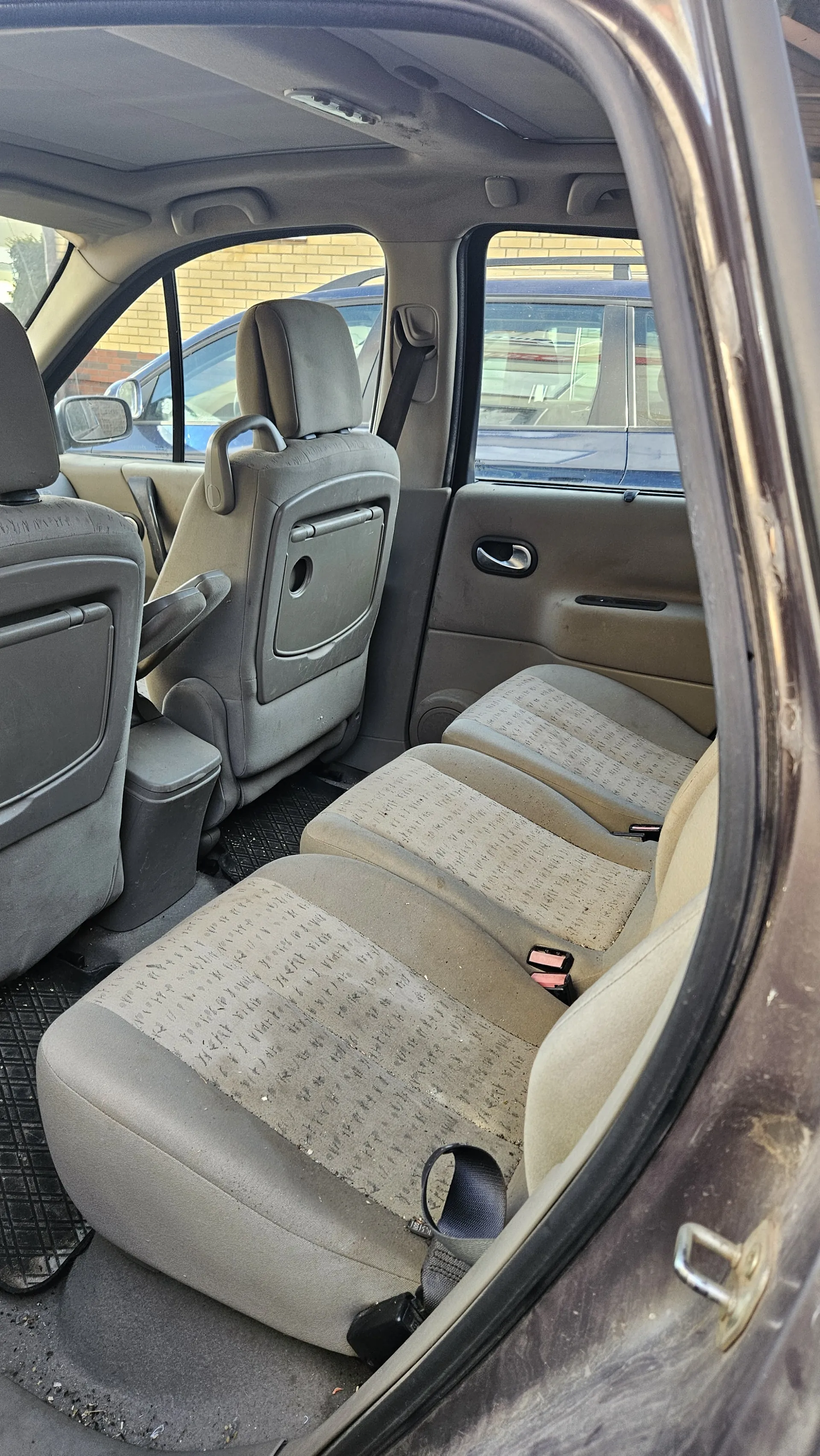 Wnętrze samochodu widziane z otwartego tylnego drzwi. Widoczne są trzy rzędy siedzeń w kolorze beżowym z wzorzystą tapicerką, częściowo zabrudzoną. Drzwi pasażerskie z uchwytem i oknem, na zewnątrz widać fragment budynku z cegły oraz inny samochód. Podłoga wyłożona czarnymi matami gumowymi. Wnętrze posiada szare elementy plastikowe, w tym oparcia siedzeń z otworami i uchwytami.