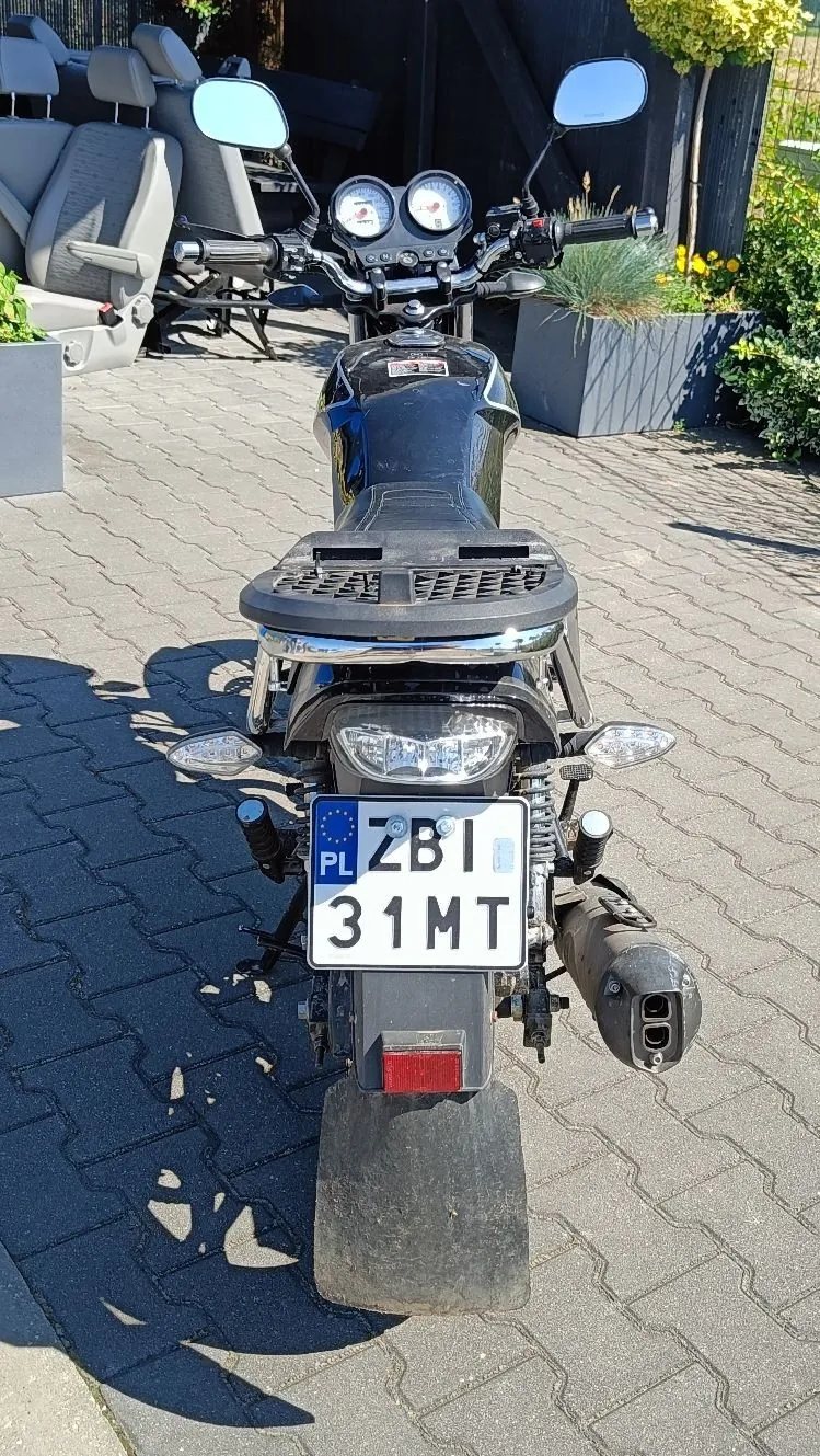 Czarny motocykl zaparkowany na kostce brukowej, widziany od tyłu. Widoczna jest tablica rejestracyjna o numerze ZBI 31MT z oznaczeniem PL. Motocykl posiada dwa lusterka, zestaw wskaźników na kierownicy oraz czarny błotnik nad tylnym kołem.