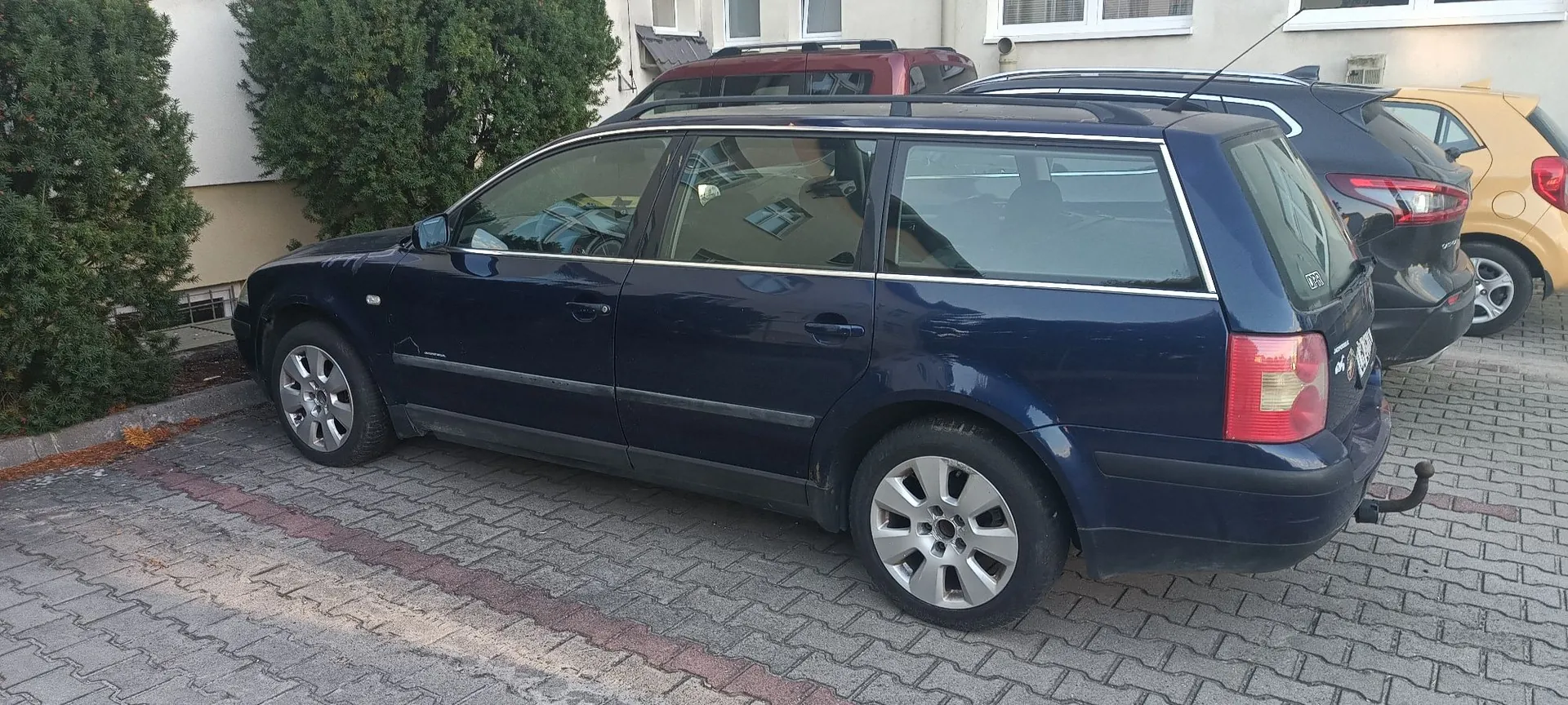 Ciemnoniebieski samochód typu kombi zaparkowany na kostce brukowej. Obok niego stoją inne samochody: czerwony i ciemnoniebieski oraz żółty. W tle widoczny jest biały budynek i zielone krzewy.