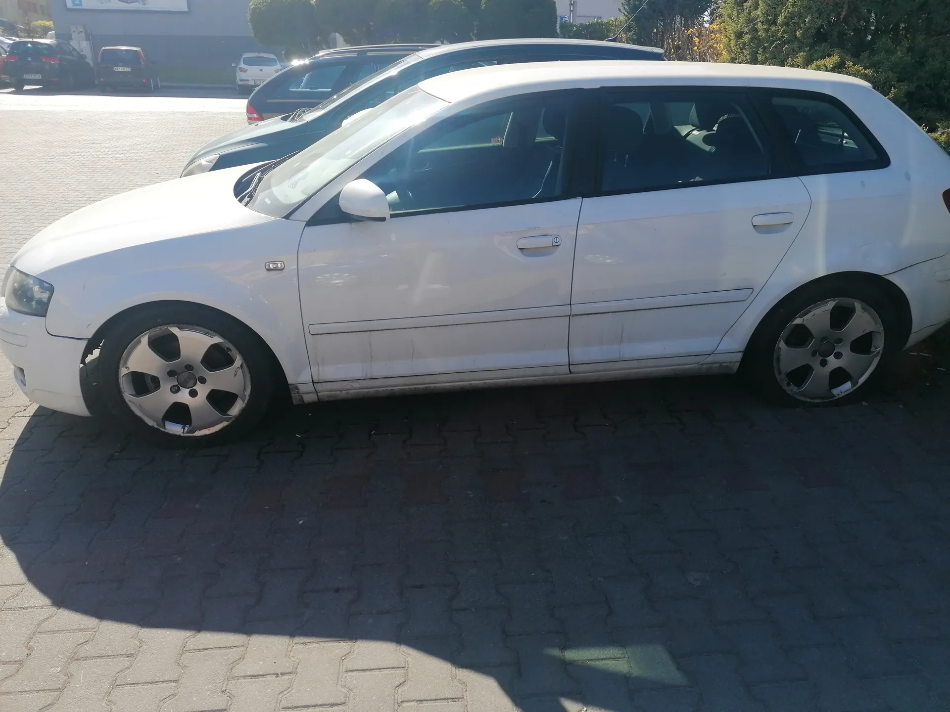 Biały samochód typu kombi zaparkowany na brukowanym placu parkingowym. W tle widoczne są inne pojazdy, drzewa oraz budynek. Samochód jest zwrócony w lewo, widok z boku. Opony i felgi są widoczne, a na boku pojazdu znajdują się ślady brudu.