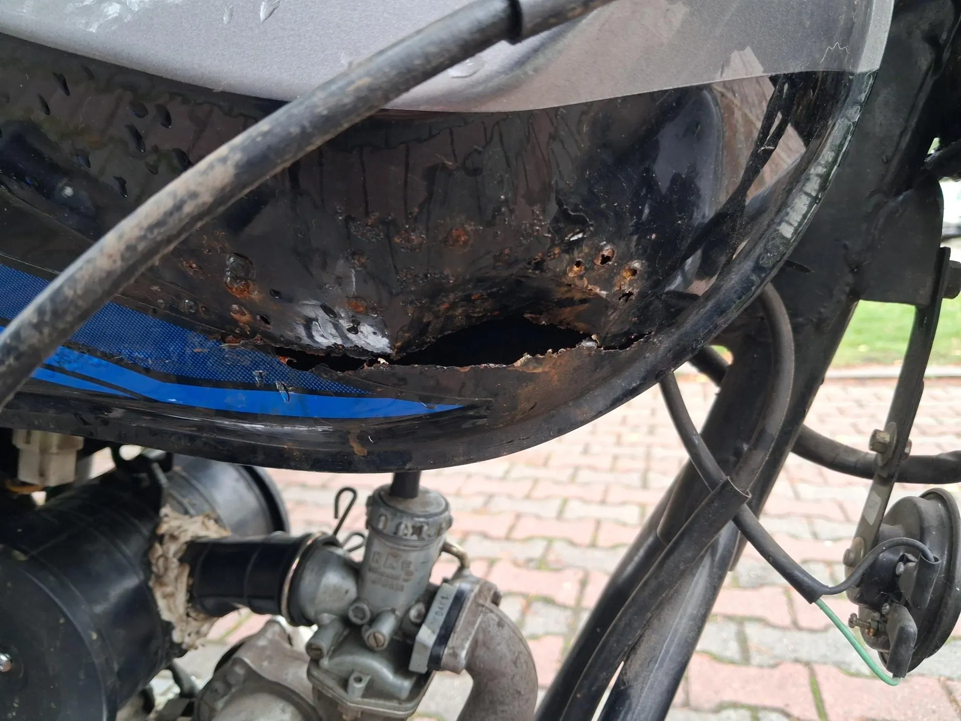 Zbliżenie na uszkodzoną, czarną metalową część pojazdu, prawdopodobnie błotnik lub obudowę, która jest silnie skorodowana z widocznymi dziurami i rdzą. Poniżej znajdują się elementy mechaniczne silnika, przewody oraz fragment kostki brukowej w tle.