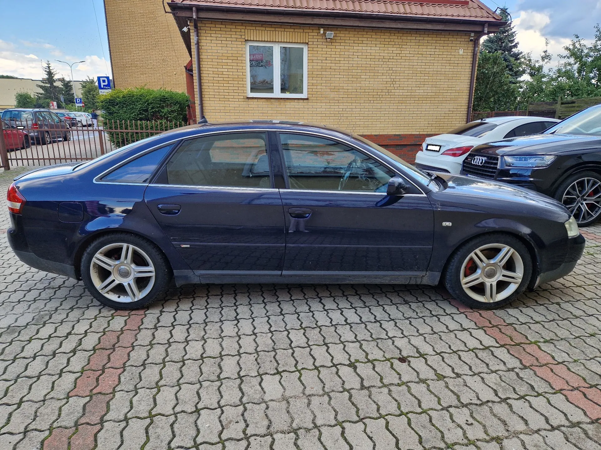 Granatowy samochód osobowy marki Audi w nadwoziu sedan, zaparkowany bokiem na kostce brukowej przed żółtym budynkiem z cegły. W tle widoczne są inne samochody oraz niebieski znak parkingowy.