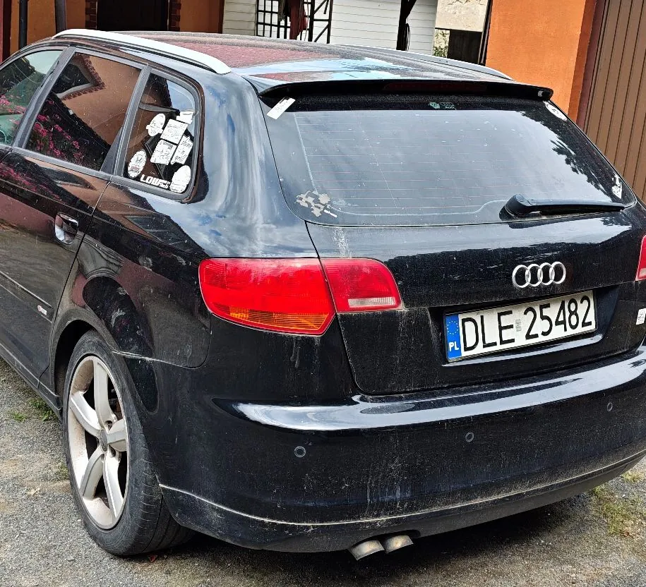 Tylny widok czarnego samochodu marki Audi w nadwoziu kombi. Pojazd jest zabrudzony, widoczna jest polska tablica rejestracyjna o numerze DLE 25482. Samochód ma srebrne felgi aluminiowe i dwie rury wydechowe po lewej stronie.
