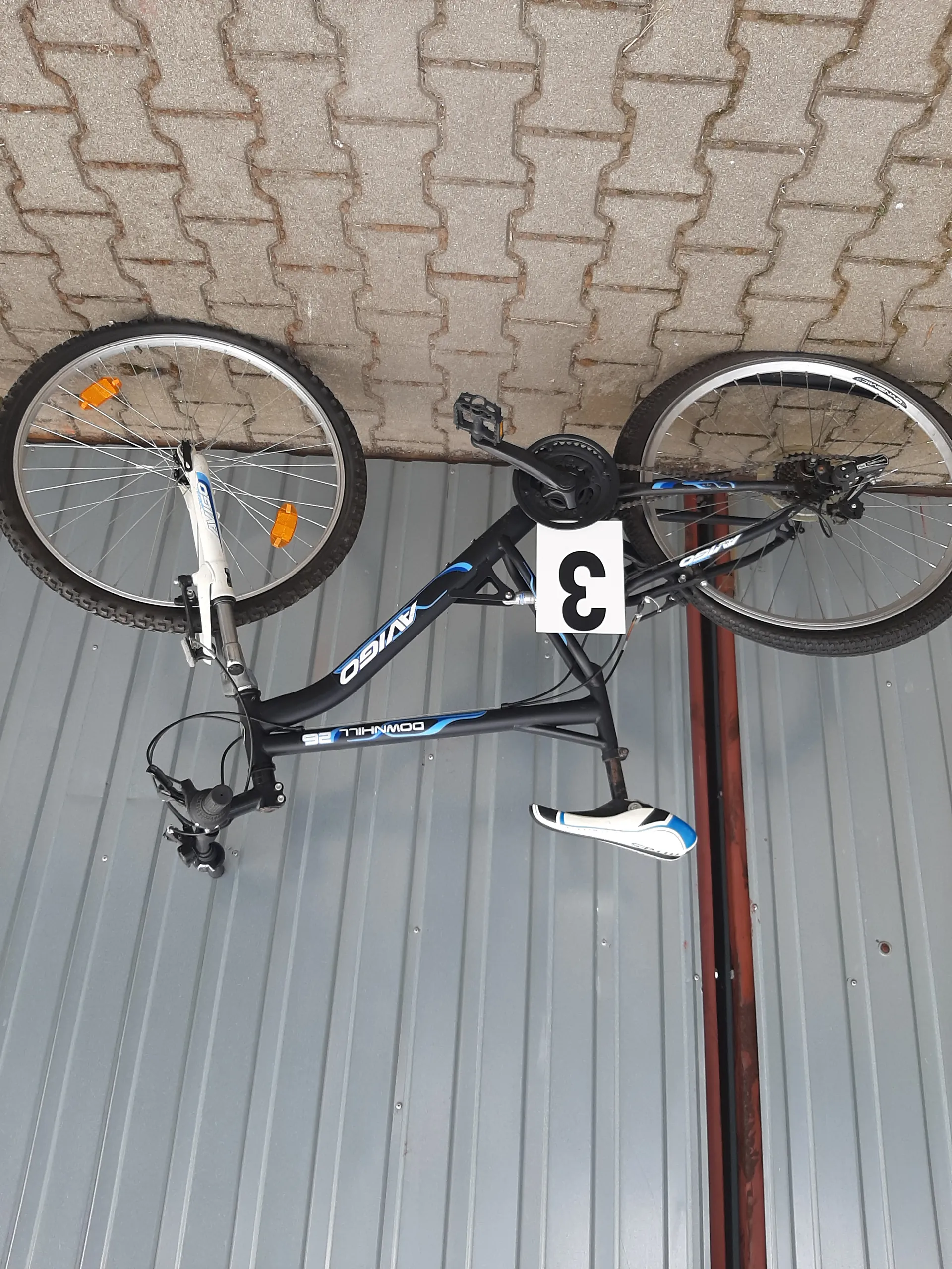 Czarny rower z niebieskimi akcentami marki AVIGO, model DOWNHILL 26, z białą tabliczką z czarną cyfrą 3 na ramie. Rower oparty o szarą metalową ścianę i brukowaną powierzchnię. Widoczne pomarańczowe reflektory na przednim kole oraz czarno-biały siedzisko.