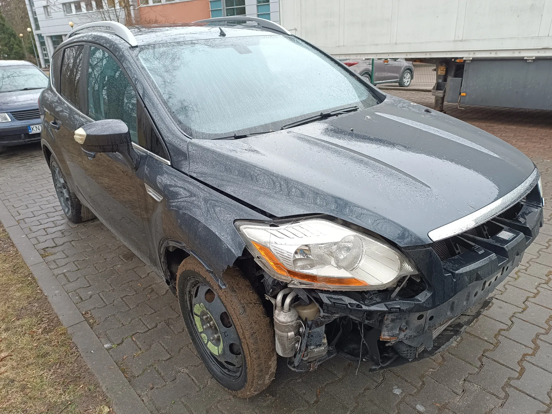 Zniszczony ciemnoszary samochód typu SUV zaparkowany na brukowanej powierzchni. Przednia część pojazdu jest uszkodzona: brakuje zderzaka, widoczne są odsłonięte elementy podwozia i części silnika. Lewe przednie koło jest zabrudzone błotem. W tle widoczny jest inny samochód z częściowo czytelną tablicą rejestracyjną KN7 oraz budynki z oknami i fragment naczepi ciężarowej. Na przedniej szybie i karoserii znajdują się krople wody.